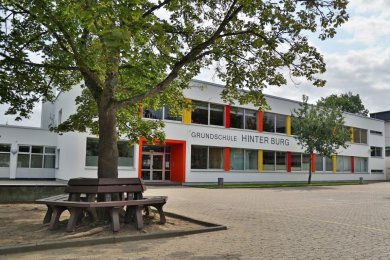 Grundschule Hinter Burg Schulhof mit einer Sitzbank rund um einen Baum, im Hintergrund befindet sich das Schulgebäude