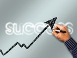 Im Hintergrund der Schriftzug "success" und im Vordergrund ein schwarzer nach oben gerichteter Pfeil der von einer Hand gezeichnet wird