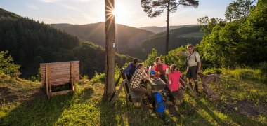 Eine Gruppe von Wanderen macht Rast am Picknickplatz Bleiberghütte. Im Hintergrund sieht man die atemberaubende Aussicht in die Wälder des Nitztals.