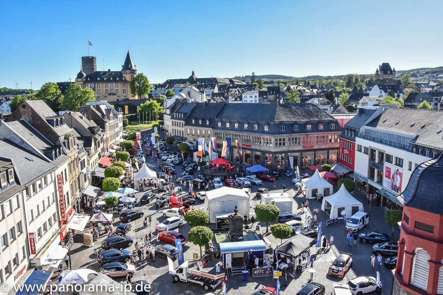 Blick aus erhöhter Position auf den Marktplatz mit Menschen und ausgestellten Autos, dazu die Genovevaburg im Hintergrund