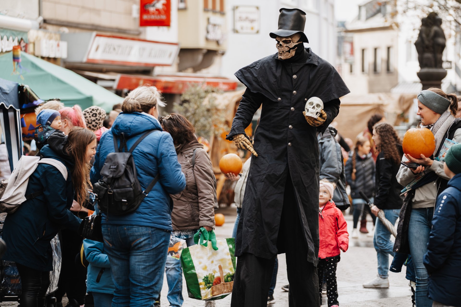 Festival der Magie ein als Tod verkleideter Stelzenläufer geht mit einem Totenkopf in der Hand durch Straße, umringt von Besucherinnen uns Besuchern