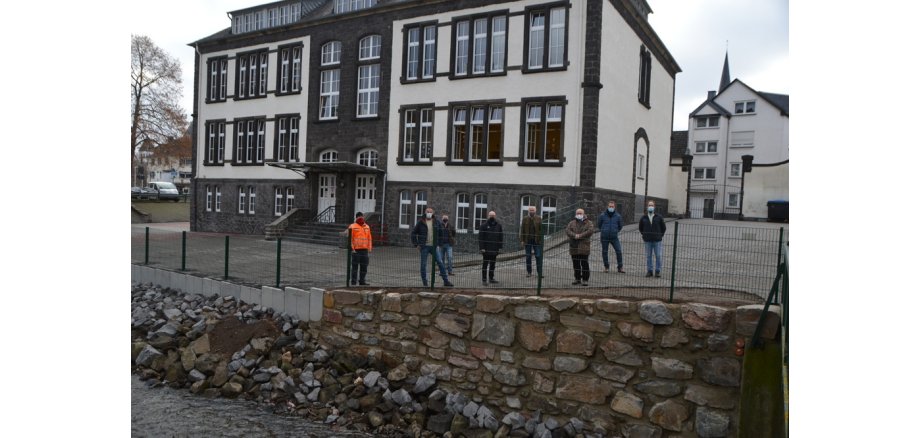 8 Personen stehen auf dem Schulhof der Clemensschule vor dem Zaun mit Blick zur Nette, das Wetter ist bewölkt. 