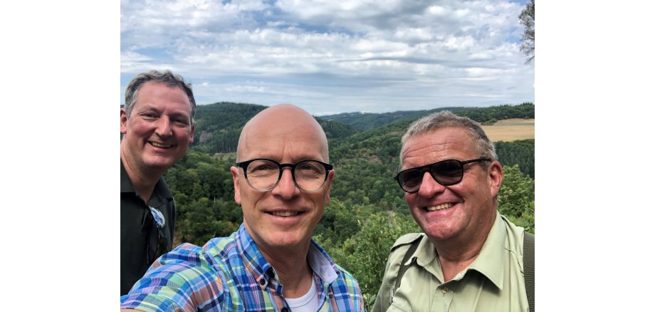 3 Personen machen ein Selfie vor einem Panorama des Mayener Stadtwaldes. Der Himmel ist bewölkt