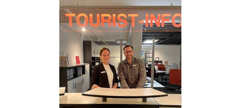 Ein Mann und eine Frau stehen hinter einer Rezeption, darüber hängt in rot das Wort "Tourist-Info"