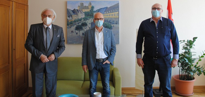 Das Bild zeigt den Hans Mayer, Dirk Meid und Hermann Wagner mit  aufgesetzten Masken und Abstand in einer Reihe im Büro des Oberbürgermeisters stehend. Im Hintergrund hängt ein Gemälde der Genovevaburg.