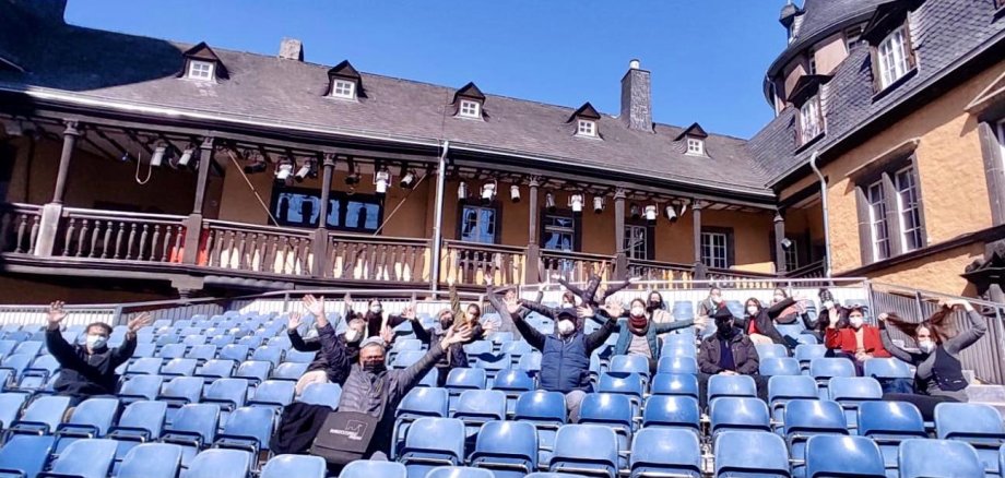 Das Ensemble der Burgfestspiele 2021 sitzt verstreut auf der Tribühne im Innenhof der Burgfestspiele. Alle tragen Masken, reißen jedoch erfreut die Arme in die Luft, weil sie unter strahlend blauem Himmel und unter strengen Hygienebestimmungen mit Proben für die Spielzeit 2021 beginnen können.