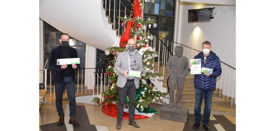 3 Männer stehen mit Gutscheinen in der Hand im Eingangsbereich der Stadtverwaltung vor dem Weihnachtsbaum mit Gutscheinen in der hand