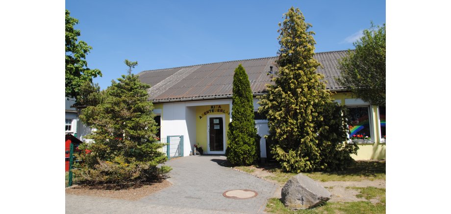 Eingangstür der Kindertagesstätte mit farbigem Schild auf dem steht KiTa Abenteuerland, rechts und links vom Eingangsweg stehen Bäume und Tannen die durch das gute Wetter und blauem Himmel schön grün wirken.