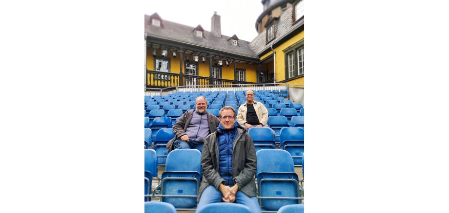 Oliver Jaksch, Charles Ripley und Matthias Manz  sitzen im Innenhof der Genovevaburg auf den blauen Stühlen mit Blick zur Bühne