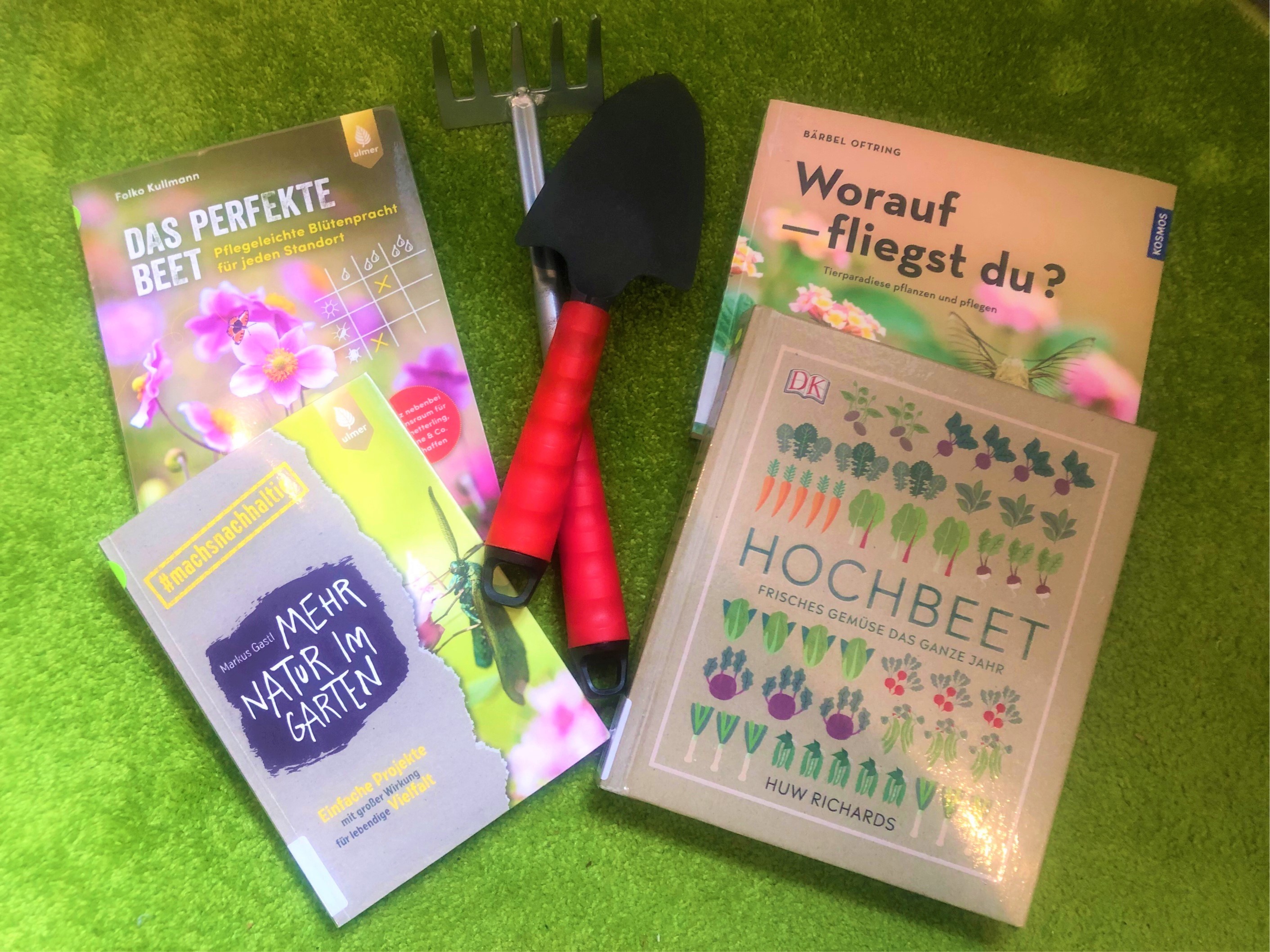 Vier Bücher zum Thema Garten, eine Schaufel und ein kleiner Rechen liegen auf einer grünen Fläche