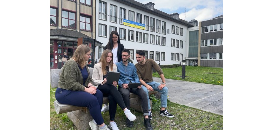 Fünf Personen recherieren vor der Verwaltung auf einer Sitzbank. Vier sitzen drauf, eine steht dahinter. 