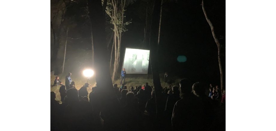 Mann steht vor einer Leinwand im Wald. Es ist dunkel. Es gibt viele Zuschauer