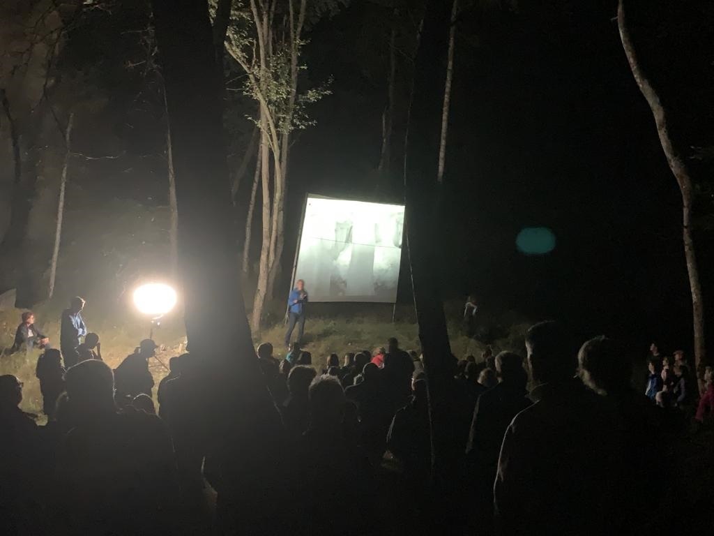 Mann steht vor einer Leinwand im Wald. Es ist dunkel. Es gibt viele Zuschauer