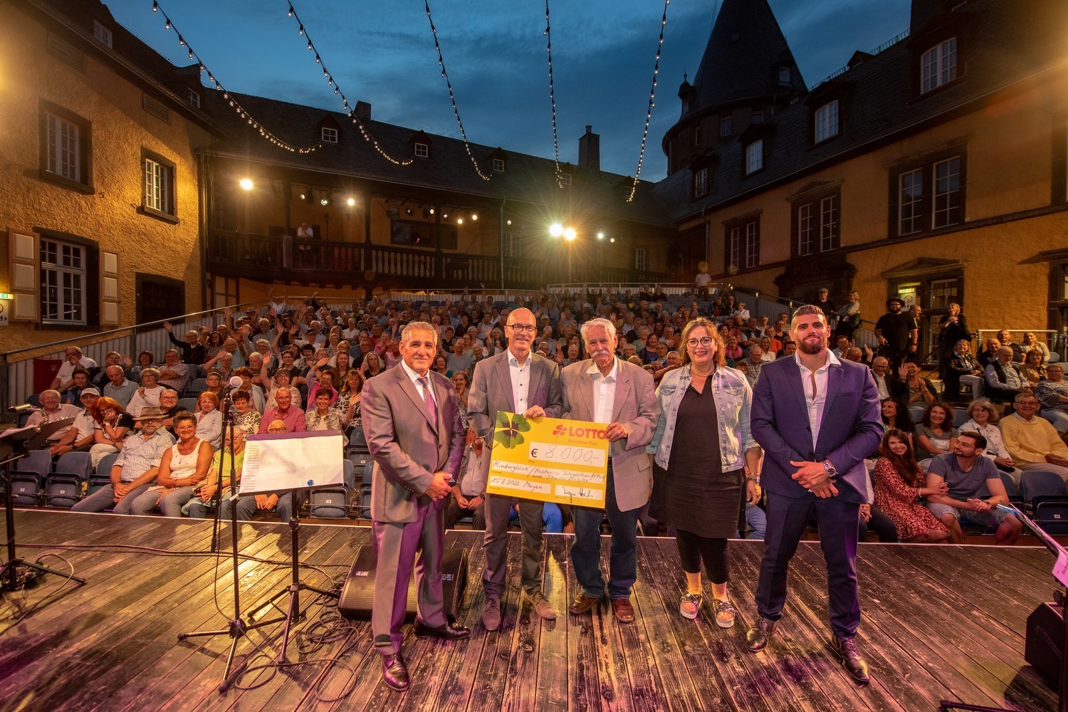 Fünf Personen stehen auf der Bühne vor dem Publikum. Zwei davon halten einen Scheck in der Hand. 