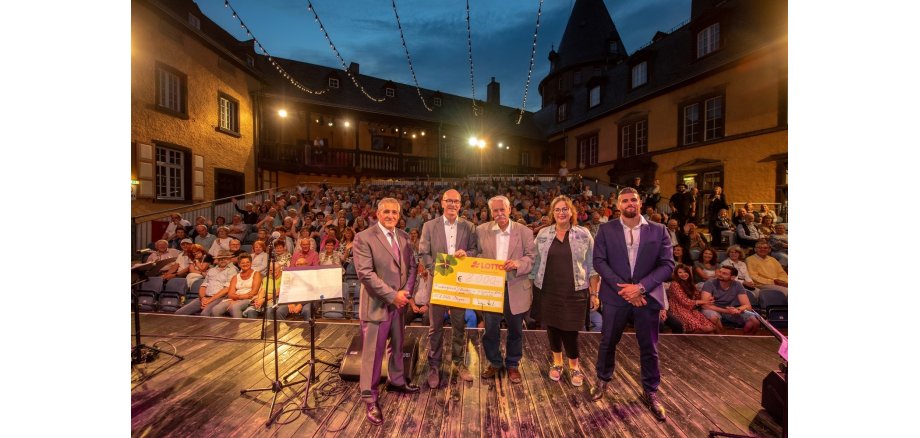 Fünf Personen stehen auf der Bühne vor dem Publikum. Zwei davon halten einen Scheck in der Hand. 