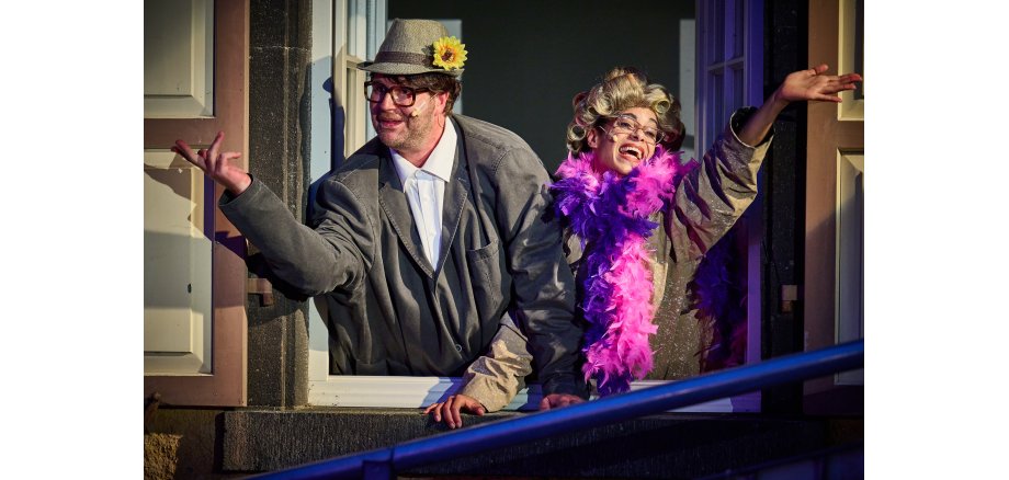 Zwei Personen stehen an einem offenen Fenster und schauen raus. Jeweils ein Arm ist rausgestreckt