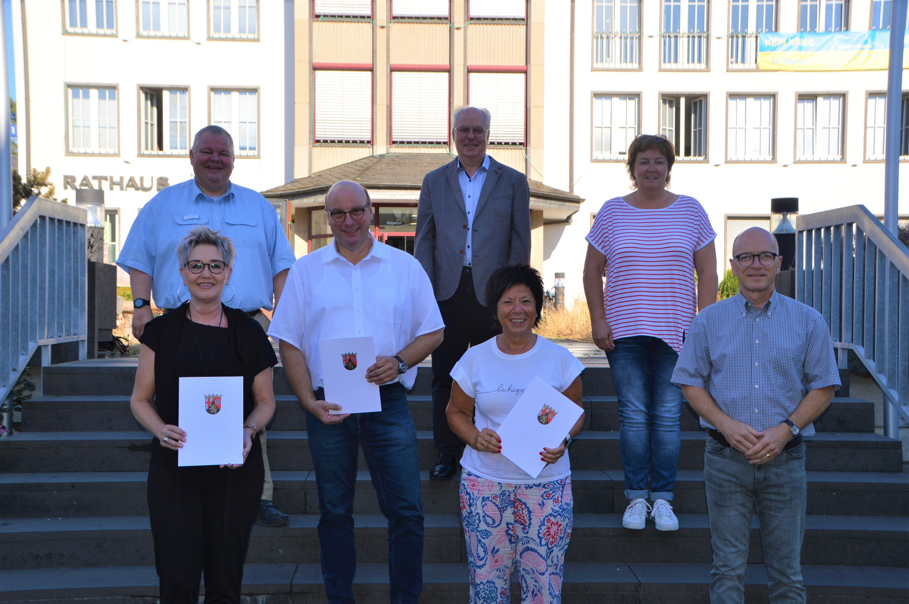 Sieben Personen stehen auf der Treppe vor dem Rathaus. Drei davon halten eine Urkunde in der Hand