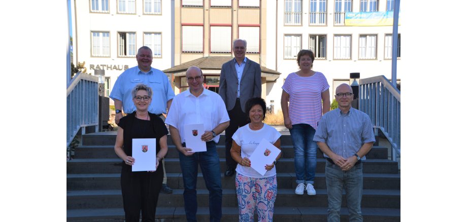 Sieben Personen stehen auf der Treppe vor dem Rathaus. Drei davon halten eine Urkunde in der Hand