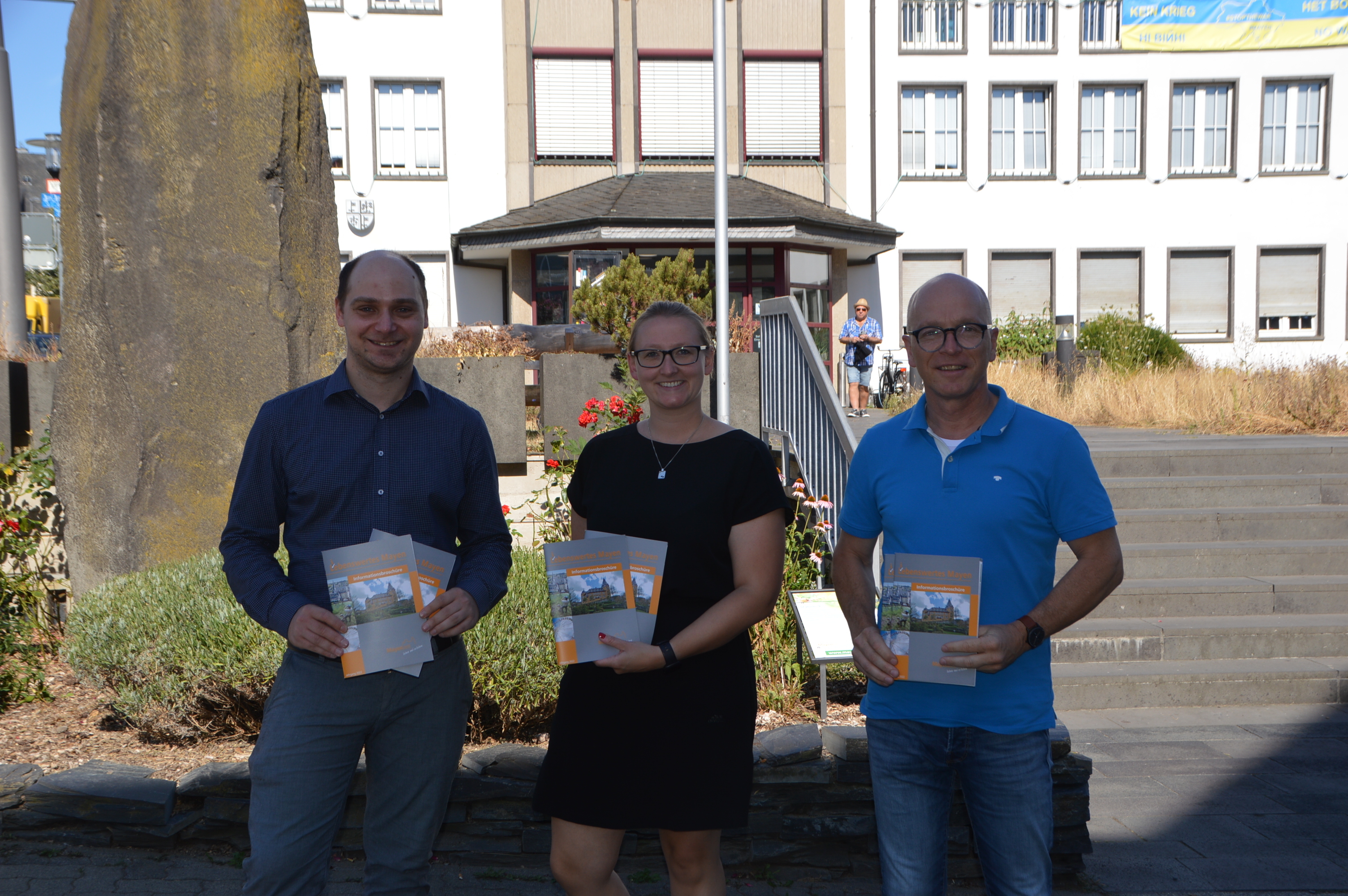 Drei Personen stehen vor der Treppe und der Basaltsäule vor dem Rathaus und halten die neue Auflage der Stadtbroschüre in den Händen