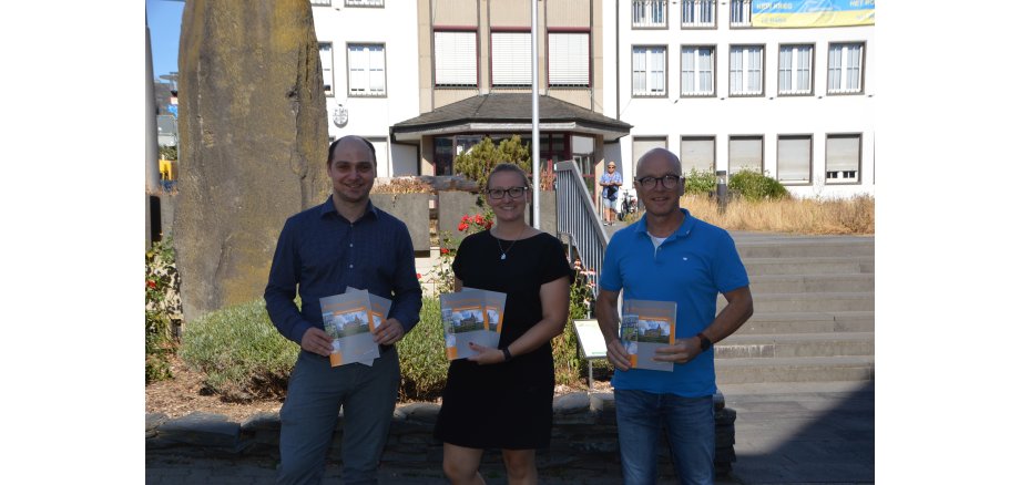 Drei Personen stehen vor der Treppe und der Basaltsäule vor dem Rathaus und halten die neue Auflage der Stadtbroschüre in den Händen