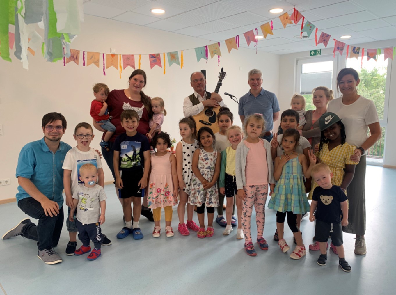 Betreuer und eine Gruppe Kinder stehen fröhlich zusammen und lächeln in die Kamera. Ein Mann hält eine Gitarre in den Händen