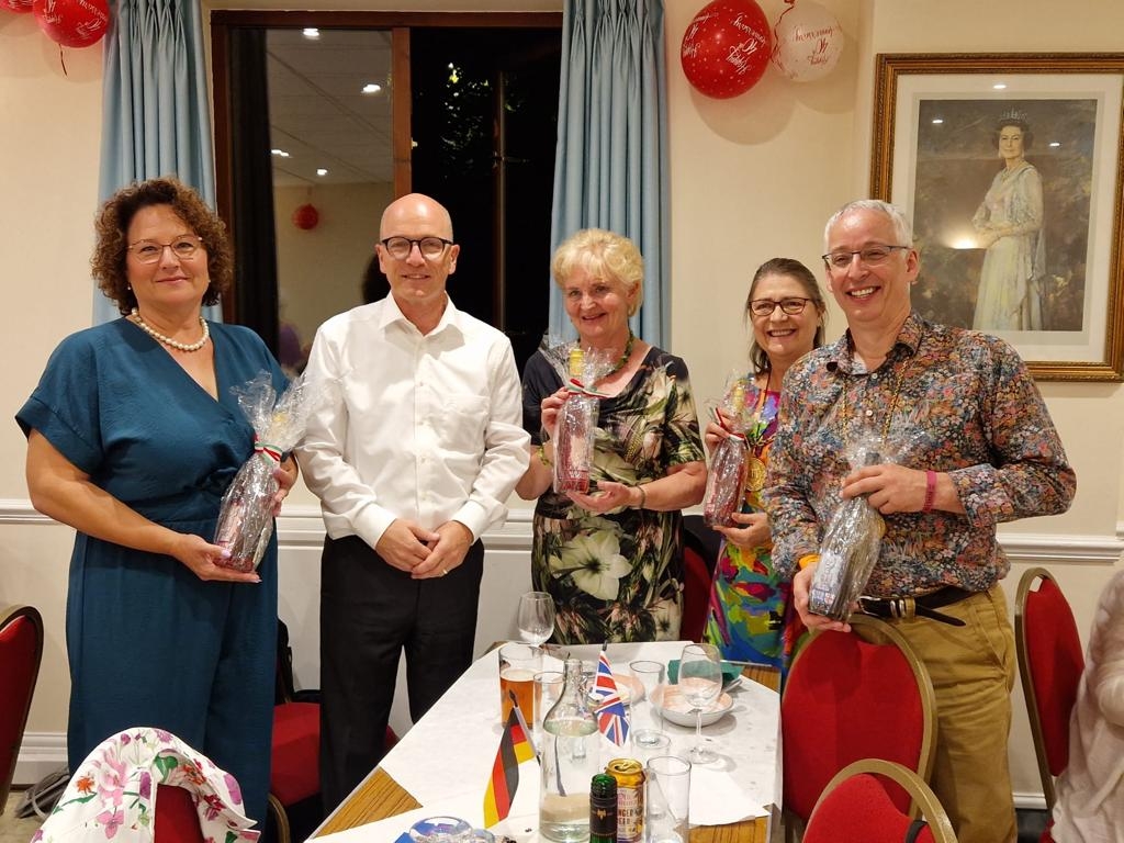 Fünf Personen stehen in einem geschmückten Raum - vier davon halten Präsente in der Hand. Alle lachen - draußen ist es dunkel