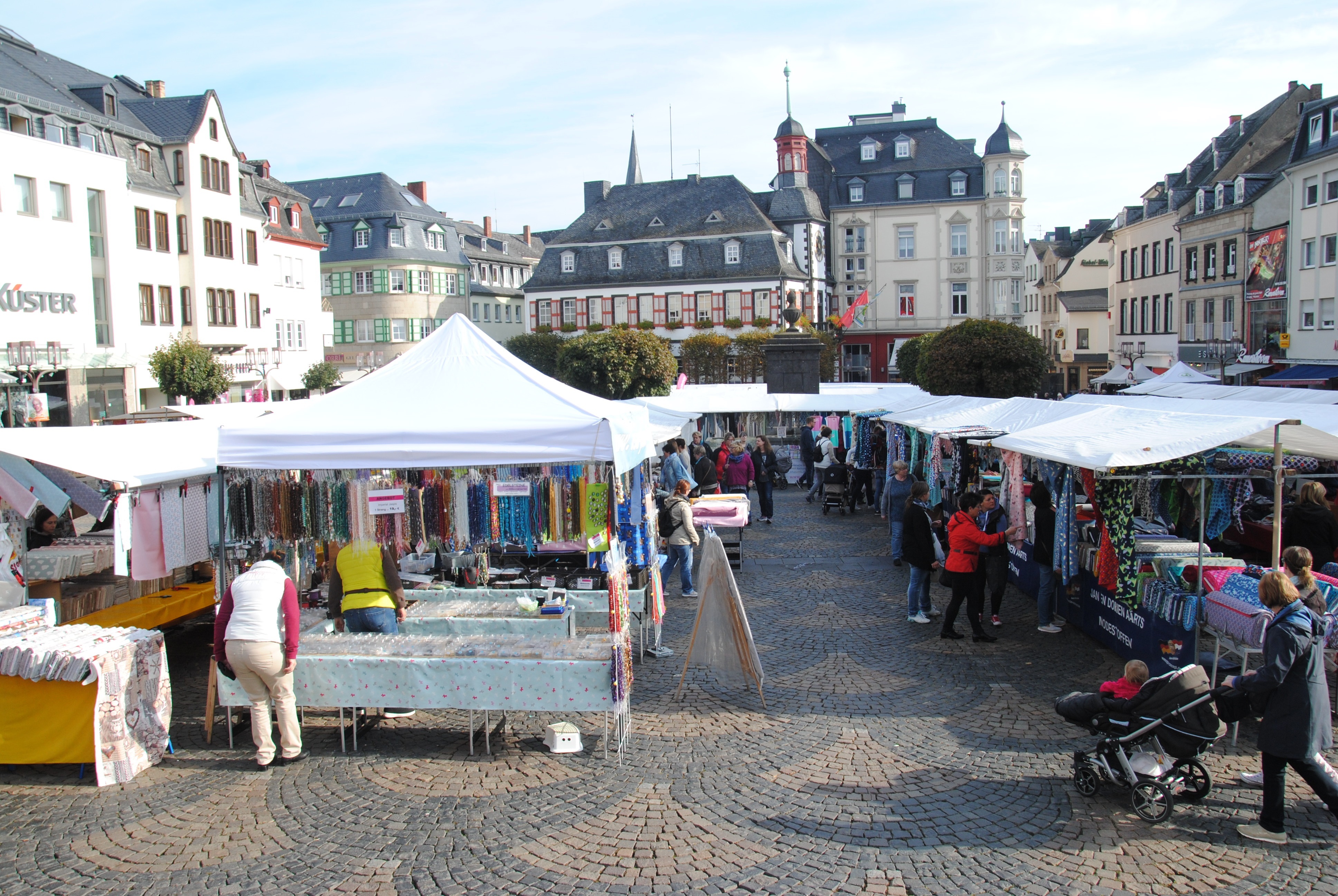Stände mit verschiedenen Stoffen und Tüchern auf dem Mayener Marktplatz. Menschen ziehen durch die Gassen. Im Hintergrund das Alte Rathaus