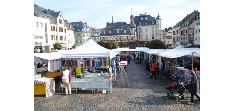 Stände mit verschiedenen Stoffen und Tüchern auf dem Mayener Marktplatz. Menschen ziehen durch die Gassen. Im Hintergrund das Alte Rathaus