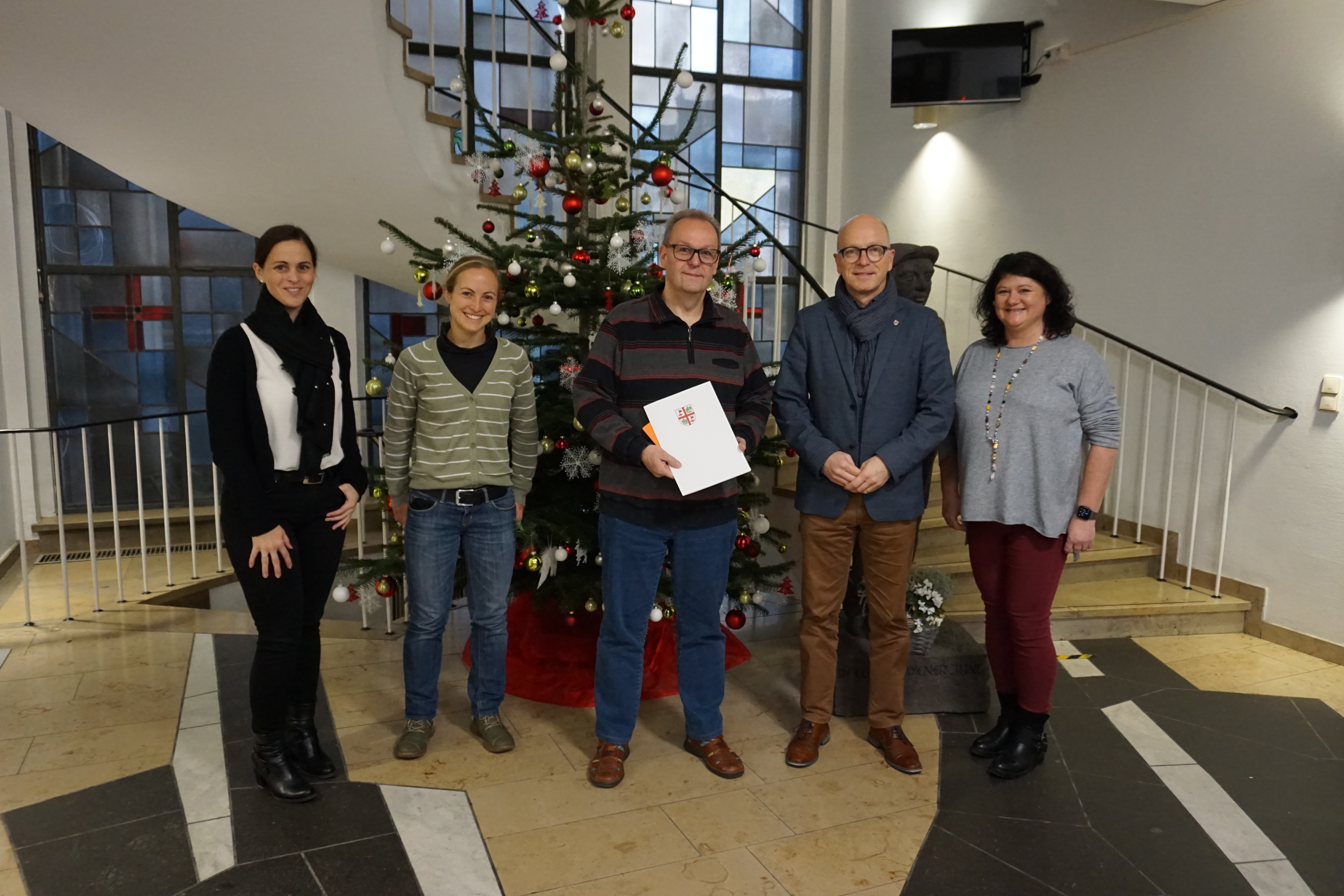Fünf Personen stehen vor einem Weihnachtsbaum im Eingangsbereich des Rathauses für ein Bild zusammen. Der Mann in der Mitte hält eine Urkunde in den Händen