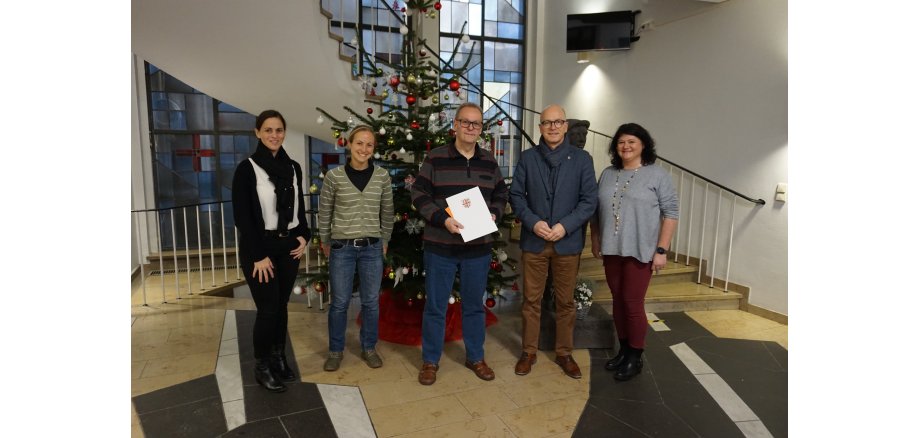 Fünf Personen stehen vor einem Weihnachtsbaum im Eingangsbereich des Rathauses für ein Bild zusammen. Der Mann in der Mitte hält eine Urkunde in den Händen