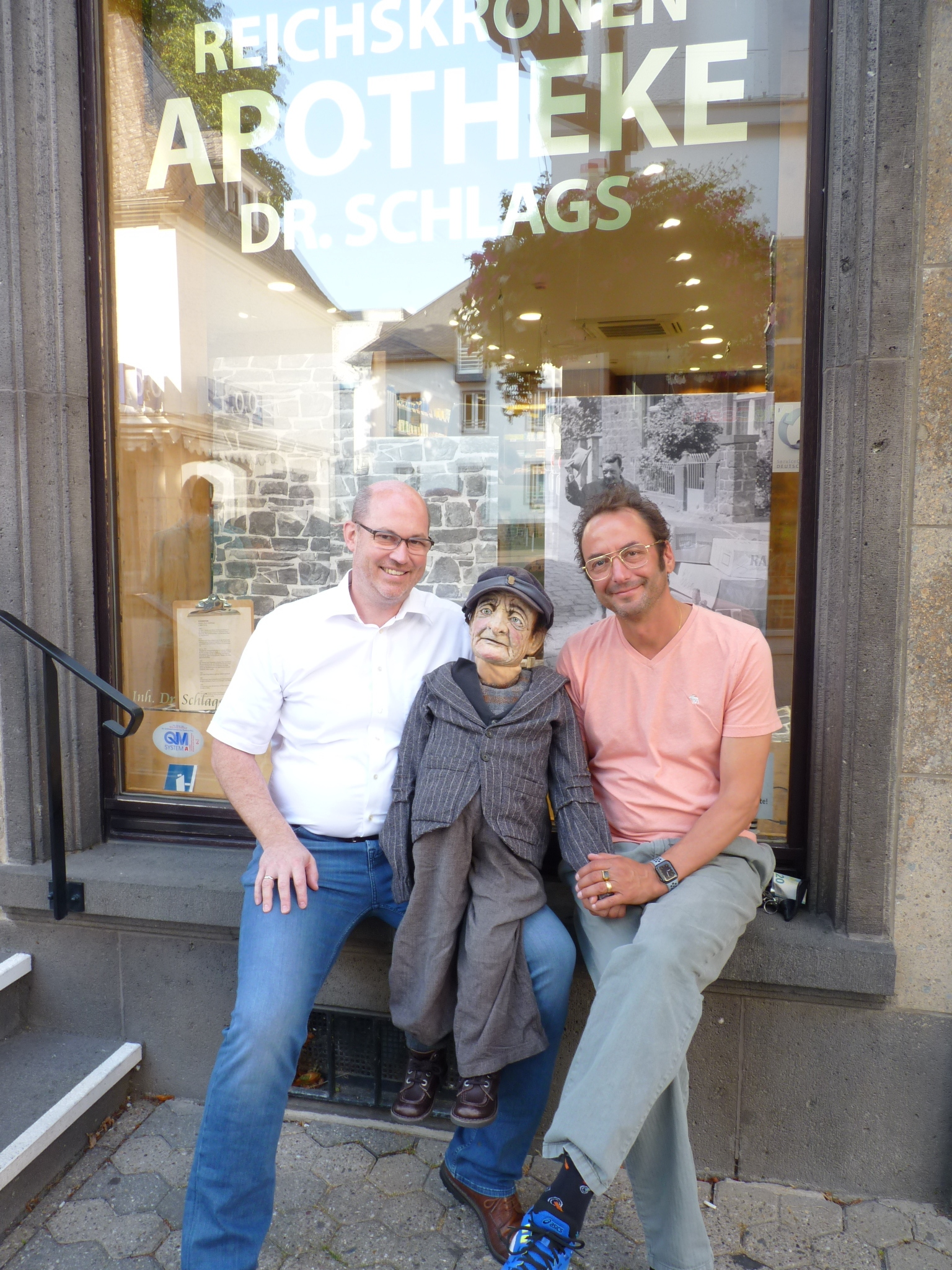 Wolfgang Schlags und Alexander May mit der Puppe auf der Fensterbank vor dem Schaufenster der Apotheke