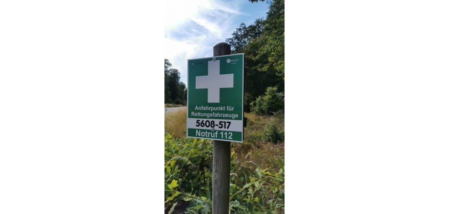 Ein grünes Schild mit einem weißen Kreuz und Notrufnummern am Waldrand