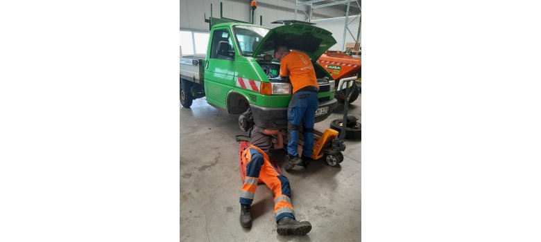 Zwei Mitarbeiter vom Betriebshof reparieren ein hochgebocktes Auto. Einer arbeitet am Motorraum und der andere liegt unter dem Fahrzeug