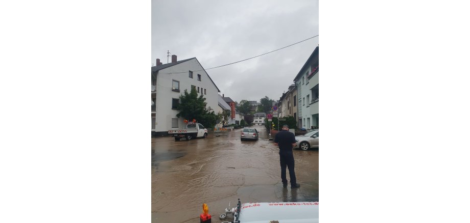 Überflutete Straße mit zwei Autos und einem Fahrzeug vom Betriebshof. Im Vordergrund steht ein Mann. 