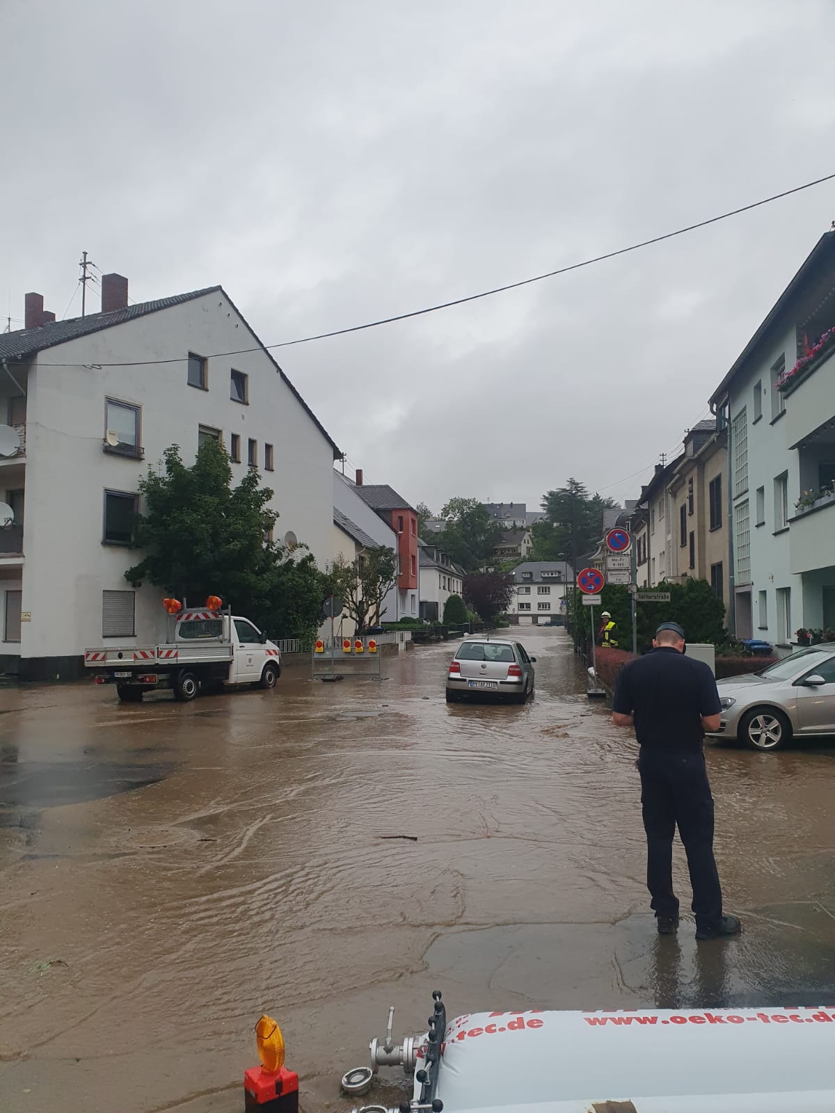 Überflutete Straße mit zwei Autos und einem Fahrzeug vom Betriebshof. Im Vordergrund steht ein Mann. 