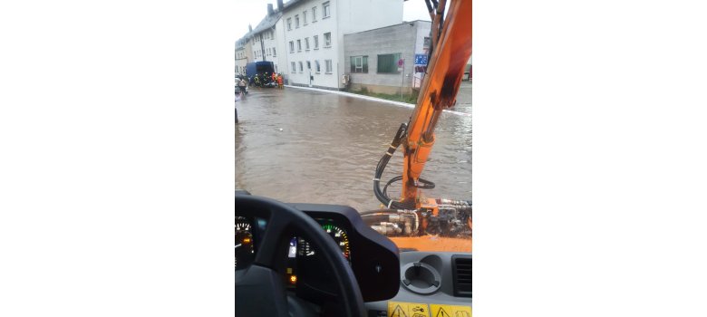 Ein Bild aus einem Fahrzeug vom Betriebshof gemacht. Das Fahrzeug steht auf einer überfluteten Straße. Im Hintergrund sieht man die Feuerwehr arbeiten