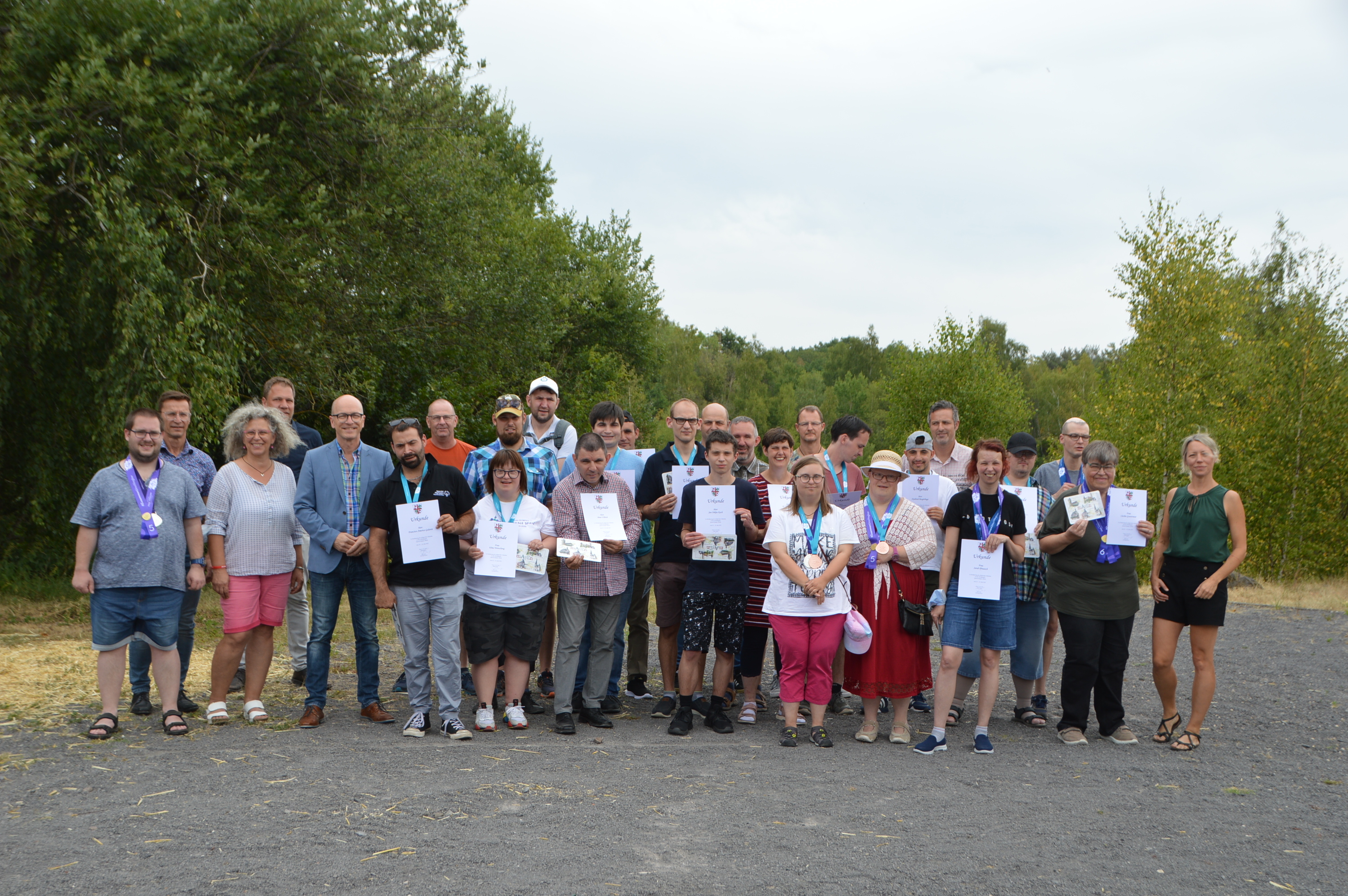 Zahlreiche Männer und Frauen - einige mit einem Dokument in der Hand stehen auf einem Platz vor Bäumen