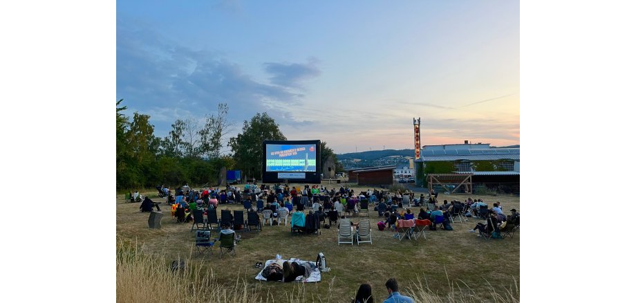 Zahlreiche Menschen mit Decken, Liegen und Stühlen sitzen auf einer Wiese vor einer großen Leinwand bei untergehender Sonne