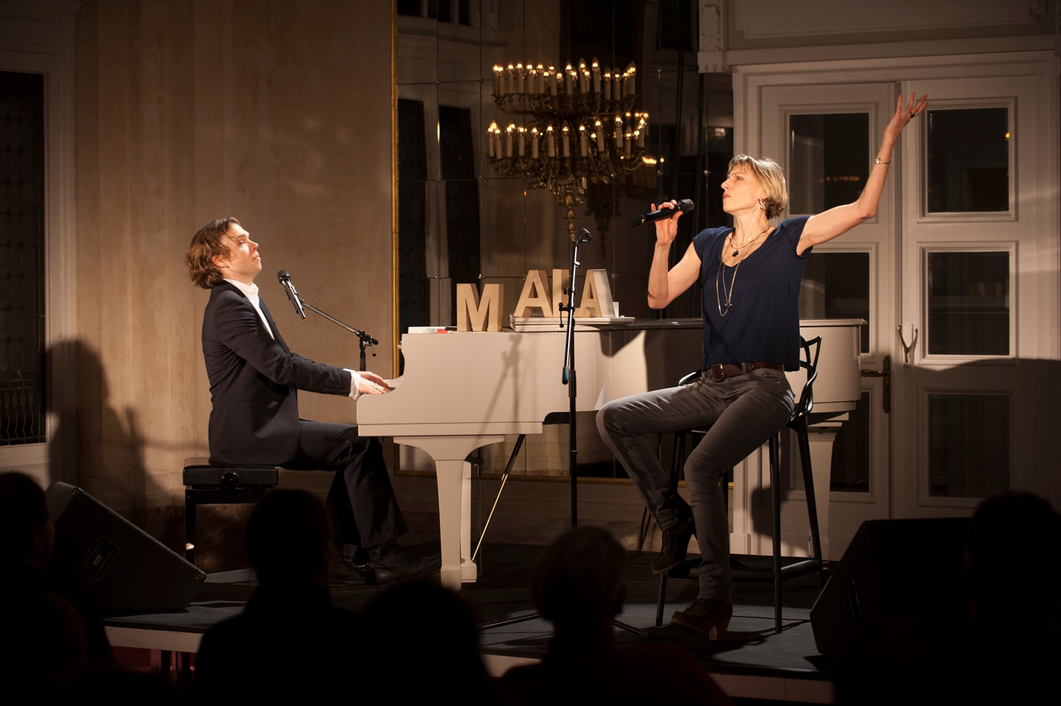 Ein Mann sitzt am Klavier in einem schwarzen Anzug und eine Frau sitzt auf einem Barkocher vor einem Mikrofonständer, hält ein Mikrofon in der Hand und hebt den linken Arm