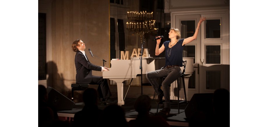Ein Mann sitzt am Klavier in einem schwarzen Anzug und eine Frau sitzt auf einem Barkocher vor einem Mikrofonständer, hält ein Mikrofon in der Hand und hebt den linken Arm