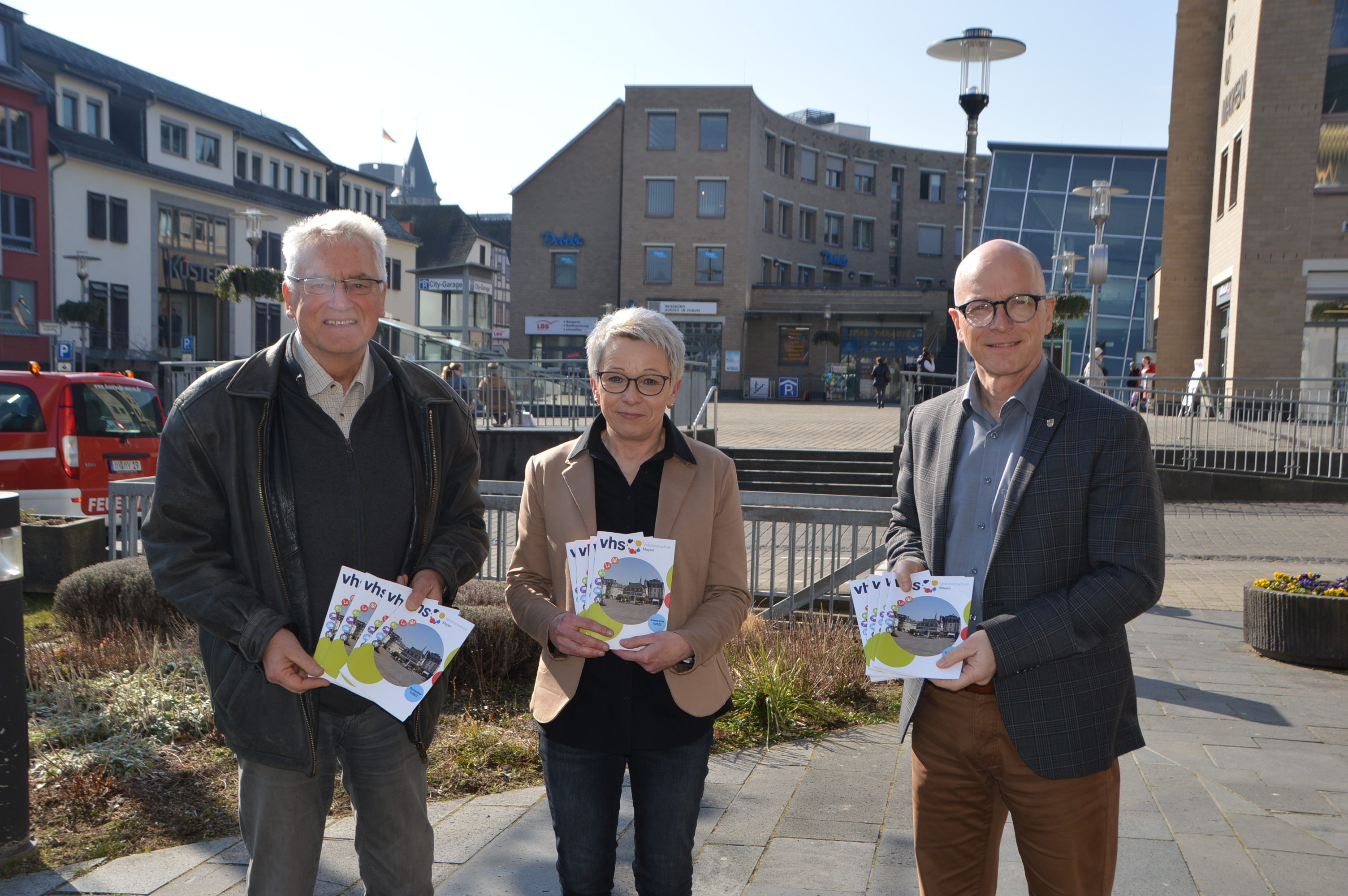 Oberbürgermeister Dirk Meid (rechts), Leona Ockenfeld von der Geschäftsstelle VHS und der pädagogische Leiter der VHS Josef Brodam präsentieren den Arbeitsplan für das I. Semester 2022 und stehen vor dem Rathaus bei schönem Wetter