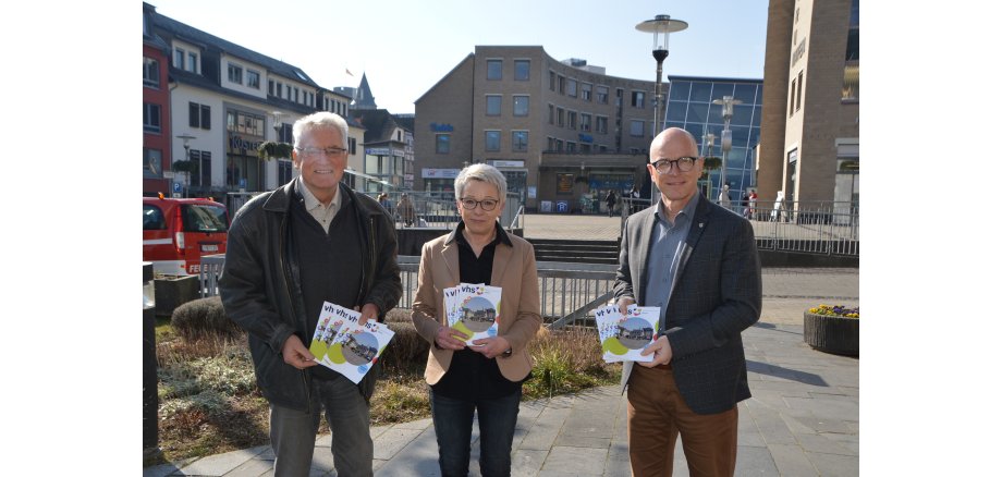 Oberbürgermeister Dirk Meid (rechts), Leona Ockenfeld von der Geschäftsstelle VHS und der pädagogische Leiter der VHS Josef Brodam präsentieren den Arbeitsplan für das I. Semester 2022 und stehen vor dem Rathaus bei schönem Wetter