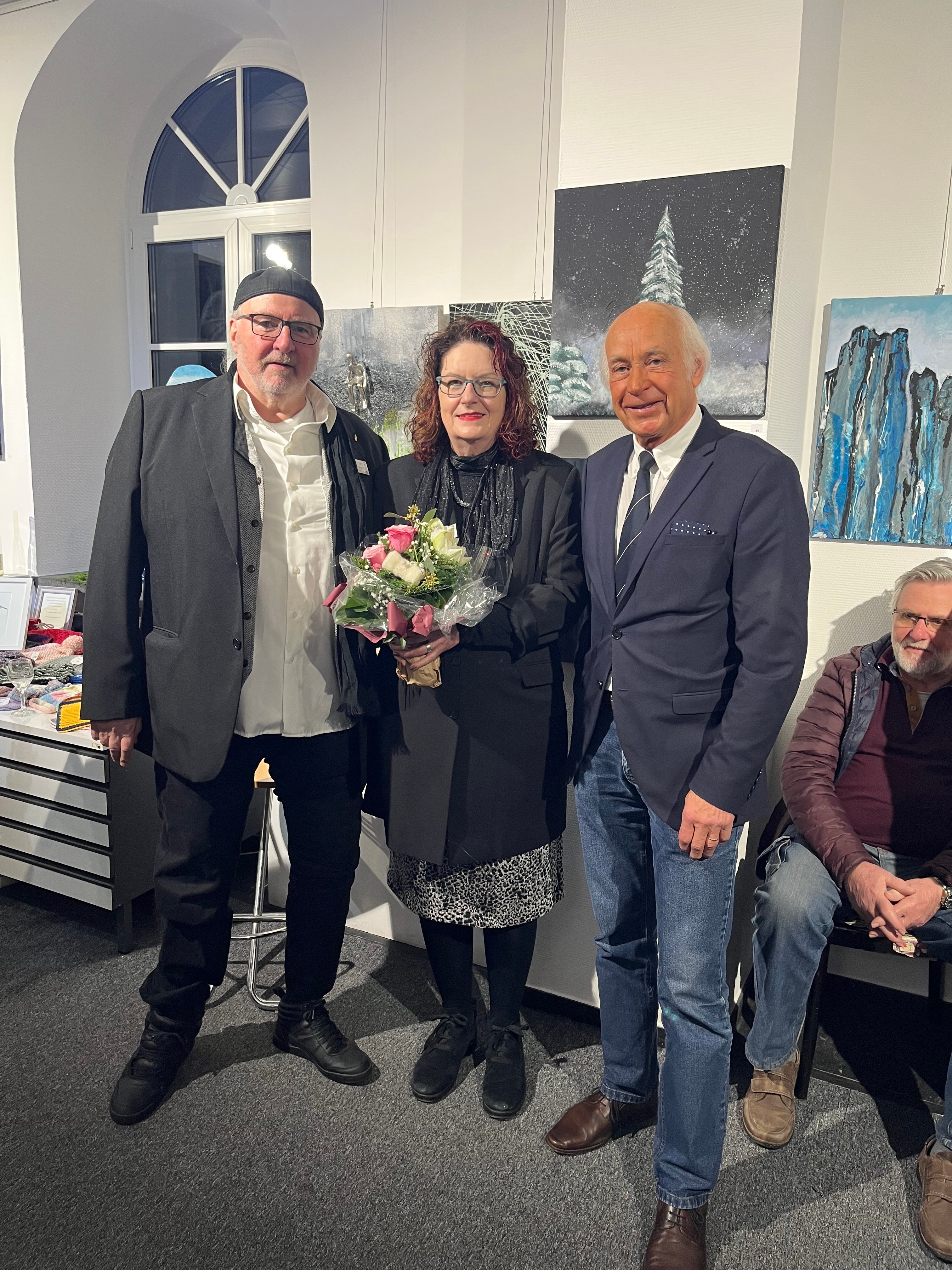 Zwei Männer und eine Frau mit Blumenstrauß stehen vor einem Gemälde