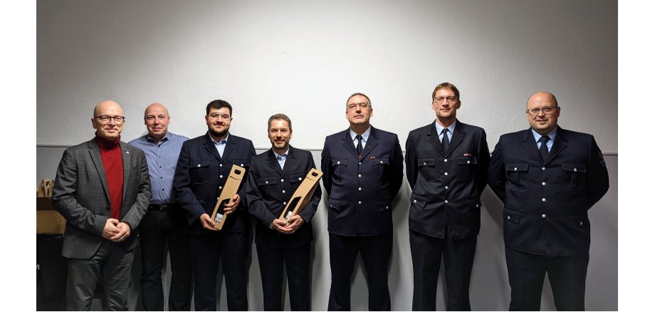 7 Personen stehen vor einer weißen Wand, 2 davon halten ein Präsent in der Hand