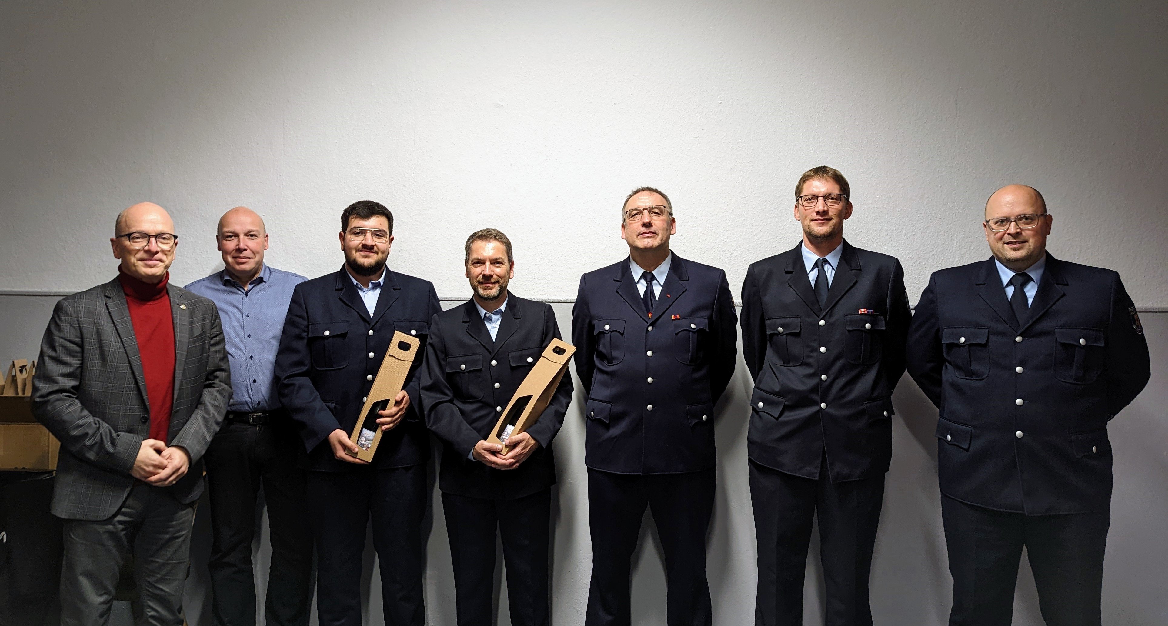 7 Personen stehen vor einer weißen Wand, 2 davon halten ein Präsent in der Hand