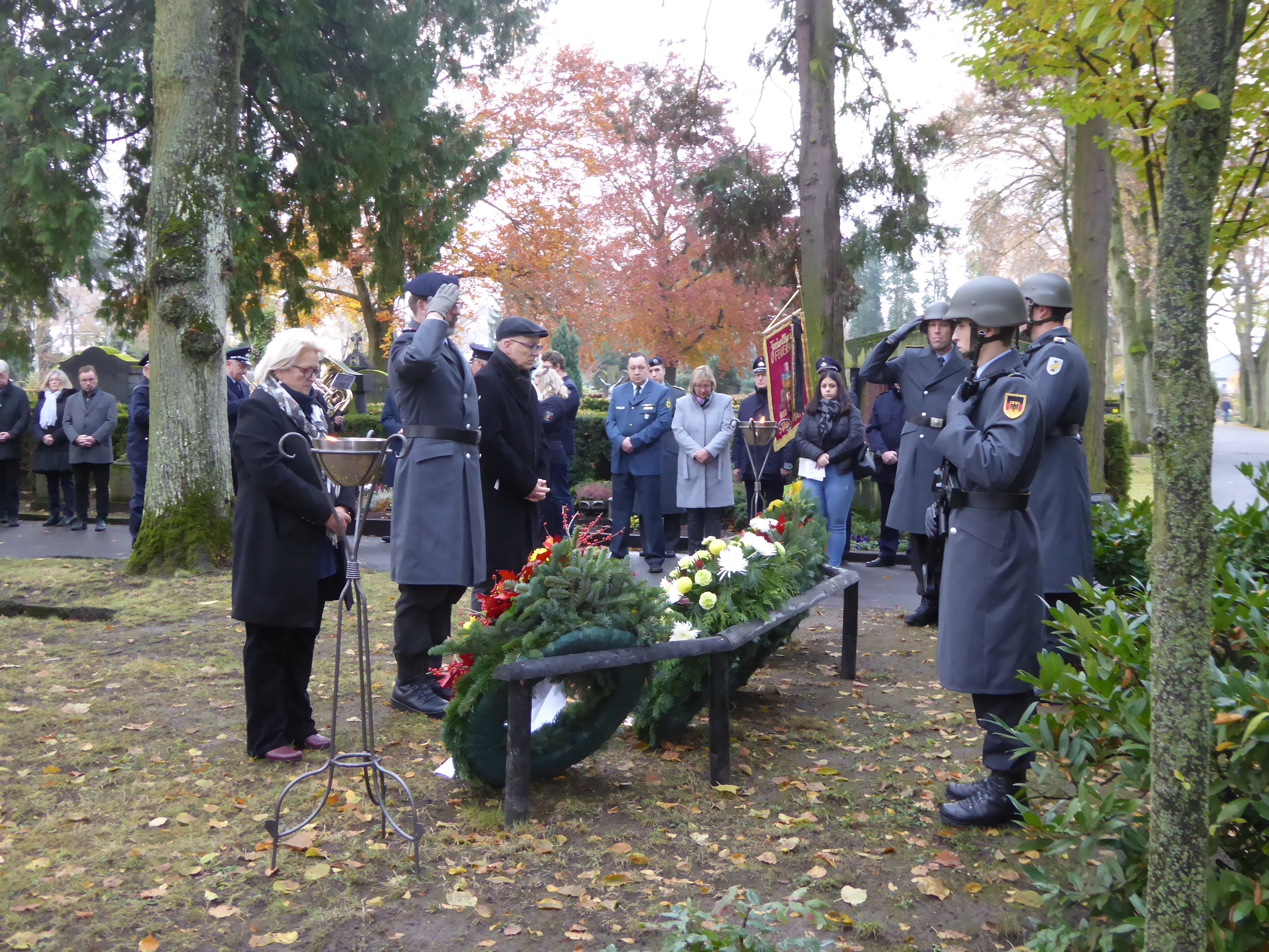 Menschen teilweise in Uniform stehen am Friedhof vor Blumenkränzen. 