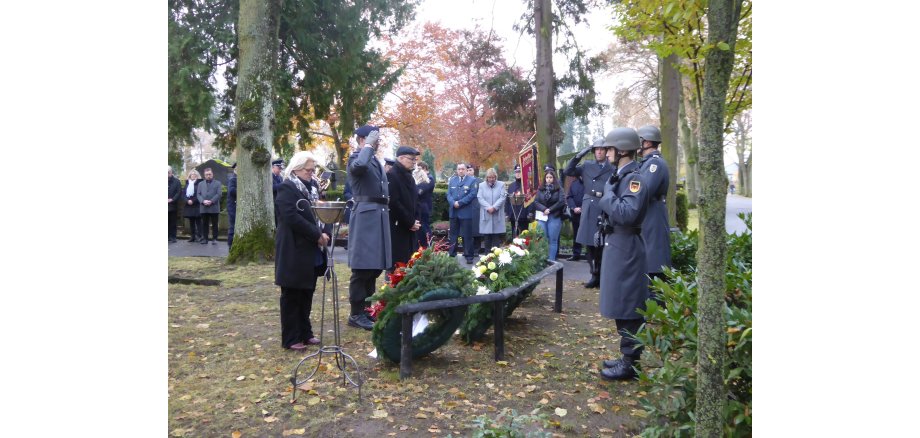 Menschen teilweise in Uniform stehen am Friedhof vor Blumenkränzen. 