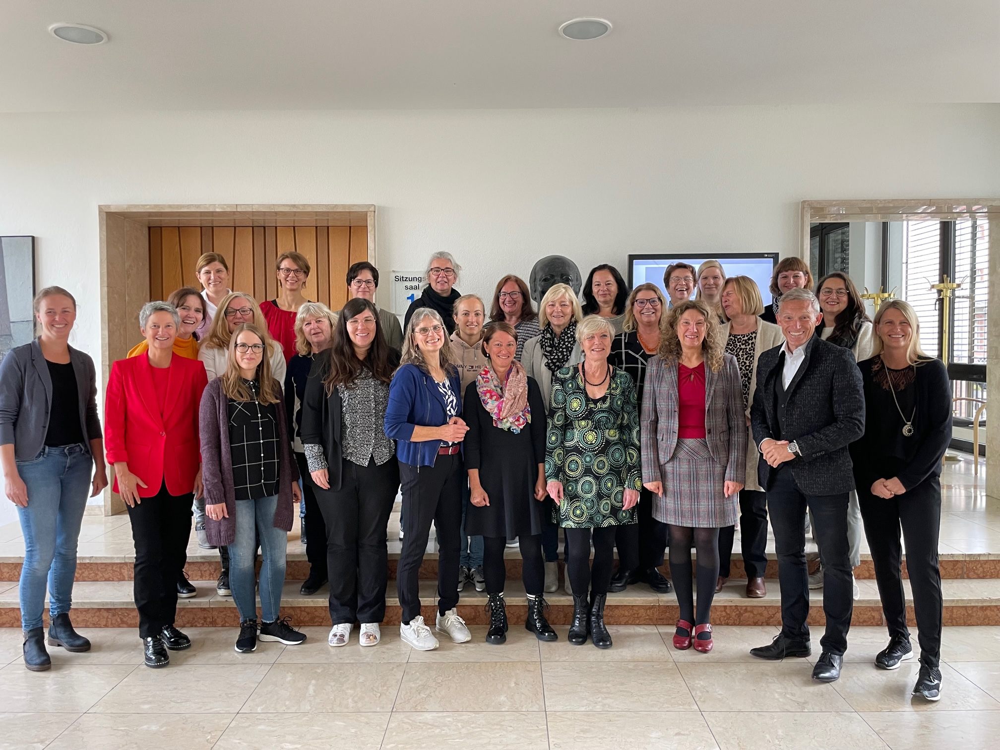 Zahlreiche Damen und ein Herr stehen in einer Gruppe verteilt auf zwei Stufen in einem Raum für ein Bild zusammen