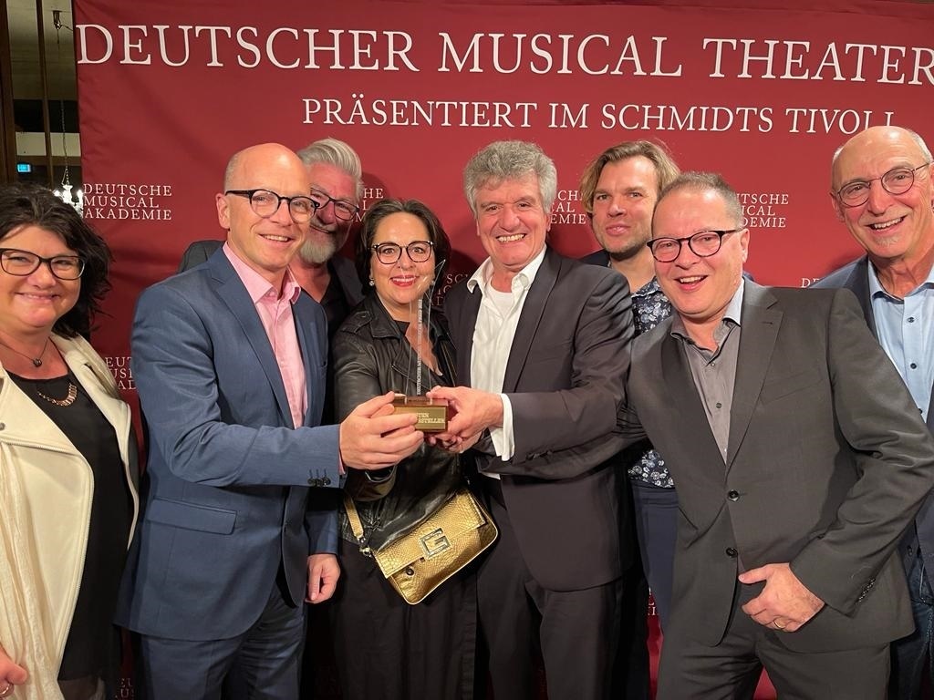 8 Personen, vor einer roten Werbewand, 3 halten den Preis in den Händen