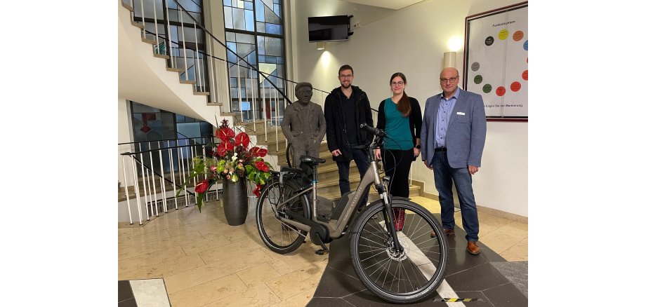 Mitarbeiter der Stadt Mayen freuen sich über Fahrrad-Leasing Zwei Männer und eine Frau stehen im Eingangsbereich der Verwaltung. Vor Ihnen steht ein E-Bike.